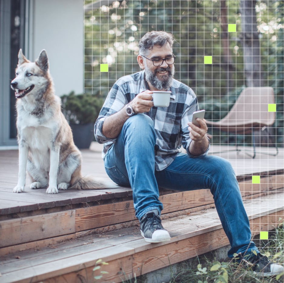 Man sitting on a backyard deck, holding a coffee mug in one hand and looking at his smartphone